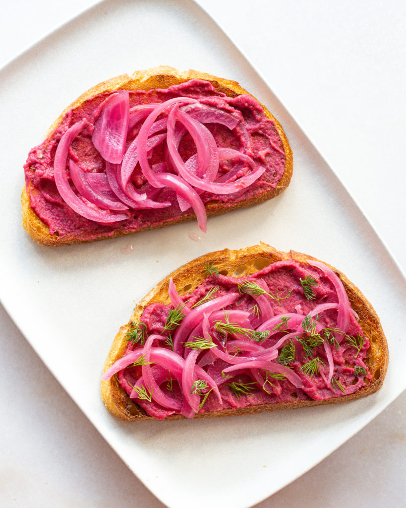 Beet hummus spread on toasted bread slices, topped with pickled red onions and dill